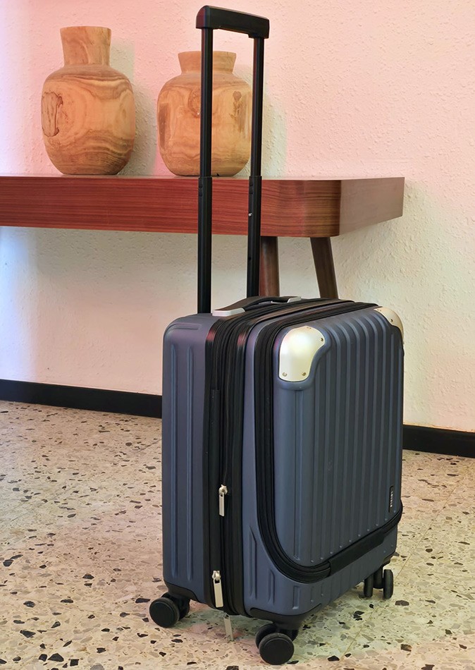 How to find the best carry-on luggage A blue carry-on suitcase standing on a gray speckled floor with a brown shelf with 2 pots in the background