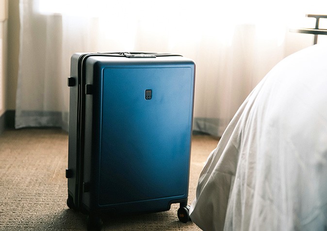 A LEVEL8 suitcase A large, sleek blue suitcase standing next to a white bed with white curtains in the background