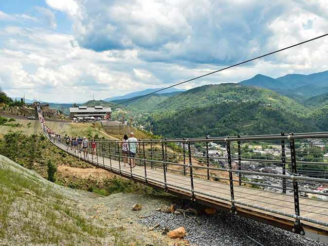 One of the best things to do in Gatlinburg A suspension bridge over a valley with green mountains and part of a town in the background