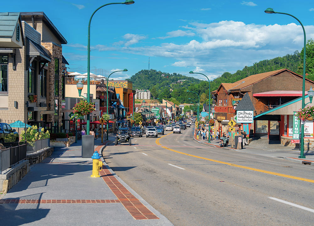 A road through f pictureque town with shops and restaurants on both sides and several people walking on the sidewalks. Green mountains in the background