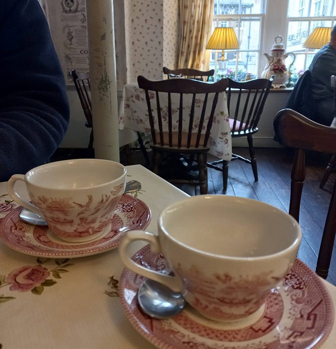 The Brits love their tea! 2 pink and white, traditional British tea cups on a table with tablecloth, with other tables and chairs and traditional British decoration in the background