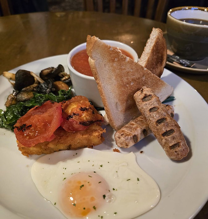 The UK is worth visiting for the food A white plate with a full English breakfast including one fried egg, 2 sausages, a hash brown, tomatoes, mushrooms, 2 slices of bread and beans in tomato sauce, plus a coffee in the background