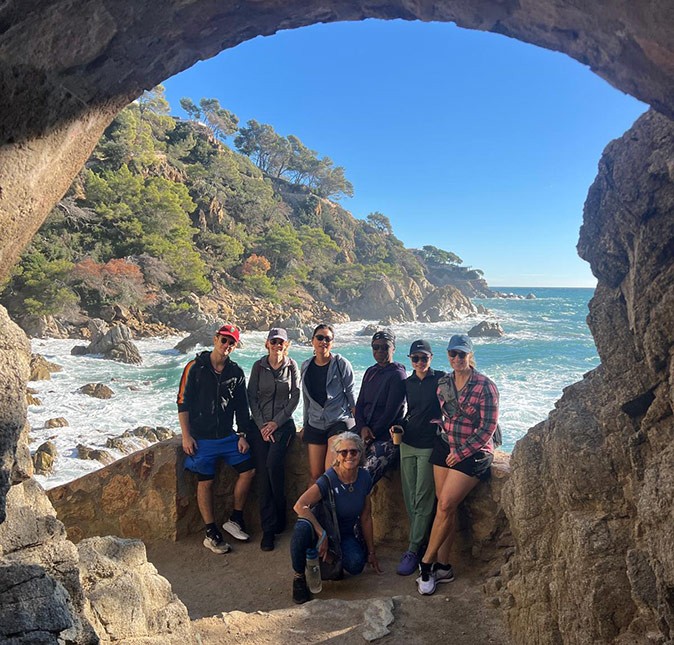 A group picture of 6 people sitting on rocks with a rocky bay, green trees and a hiking path in the background