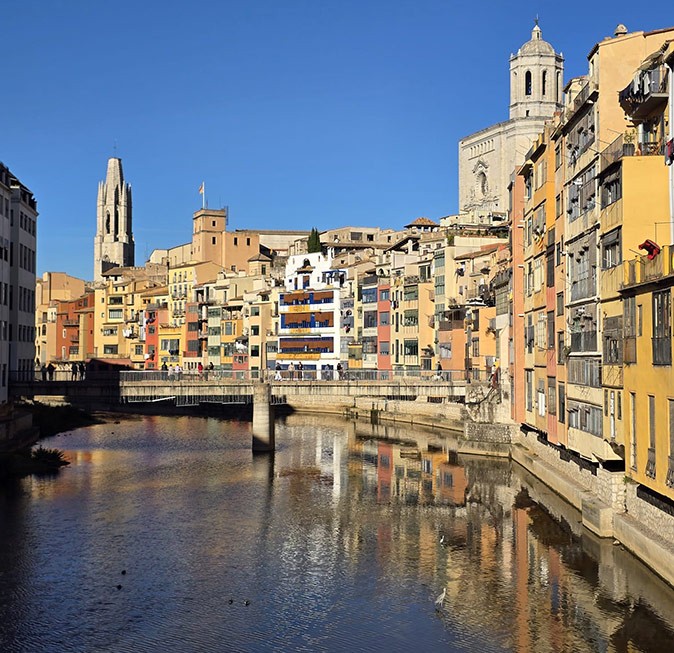 A river in the middle of a city, showing a beige bridge and beige, yellow and pink buildings on one side of the river