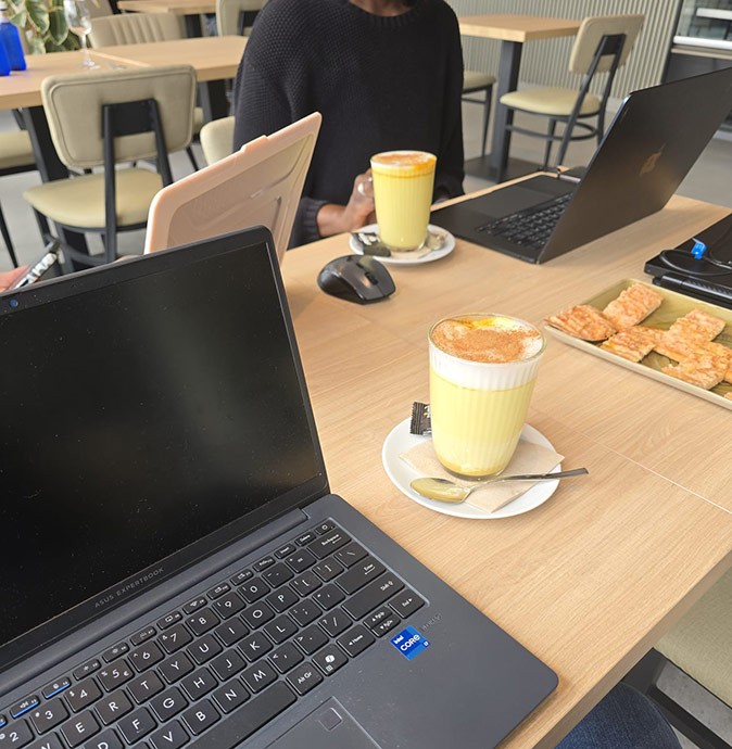 3 laptops and 2 turmeric lattes on a wooden table with parts of other tables showing in the background