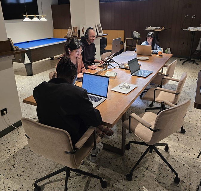 4 people sitting in office chairs around a long wooden table working on their laptops