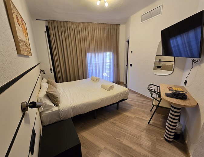 A hotel room with a white double bed, a small desk with a chair and a tv on the wall