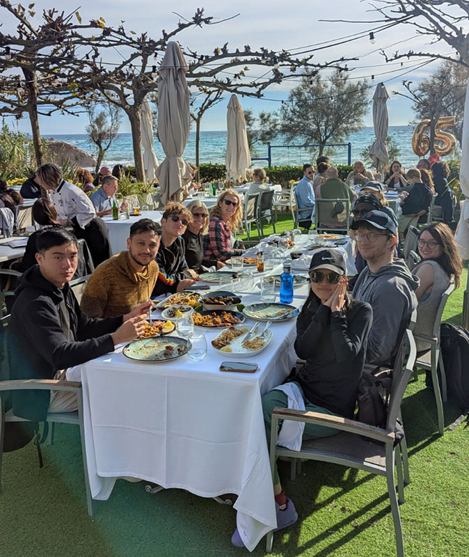 A large group of people sitting at a table with a white tablecloth, outdoors on a terrace with a green floor, some trees and the sea in the background