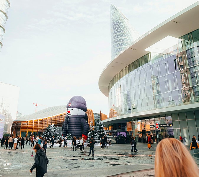A modern city square with a large glass building, several people and a large inflatable snowman