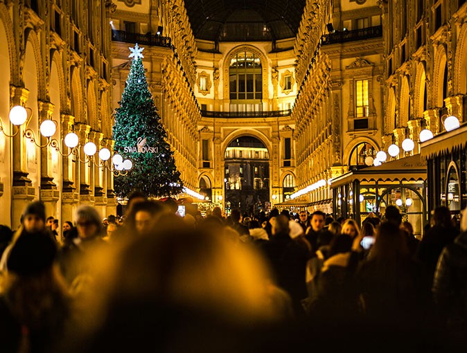 A crowd of people walking, at night, with brown buildings on both sides, atmospheric lights and a large Christmas tree