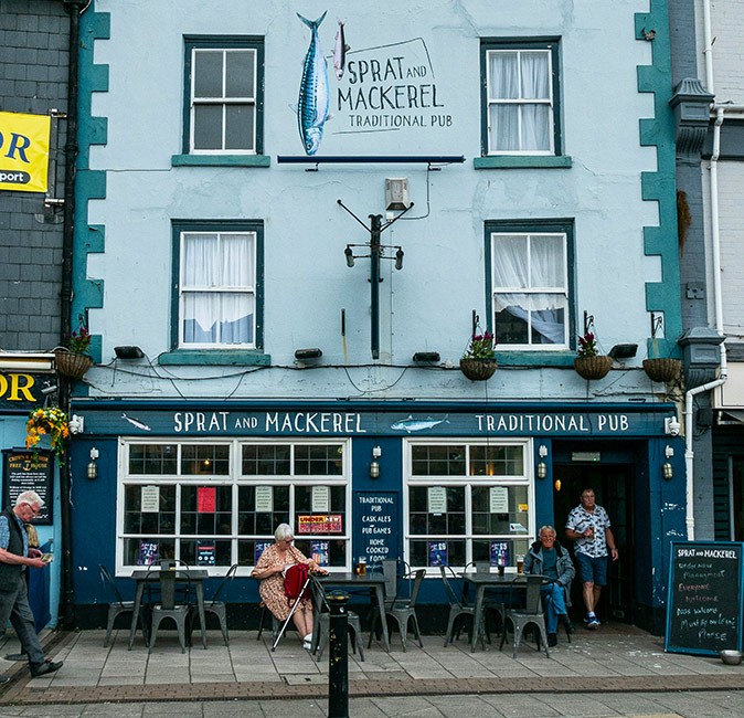 Visit a British Pub when you're in the UK The exterior of a traditional British pub, painted in shades of blue and green, with people sitting at small outdoor tables