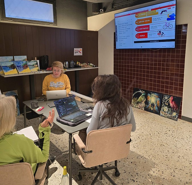 3 people sitting around a gray table, with a tv screen in the background showing Spanish grammar