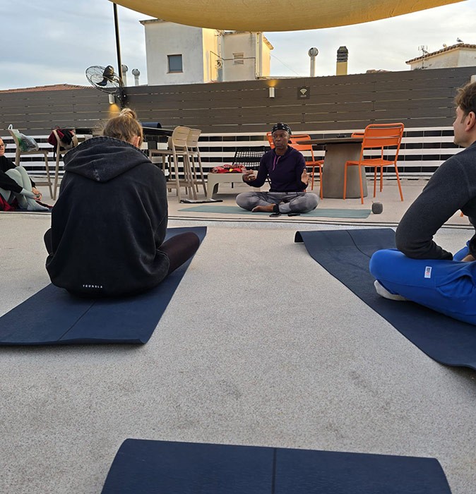 3 people sitting on yoga mats on a roof terrace with bright orange chairs in the background