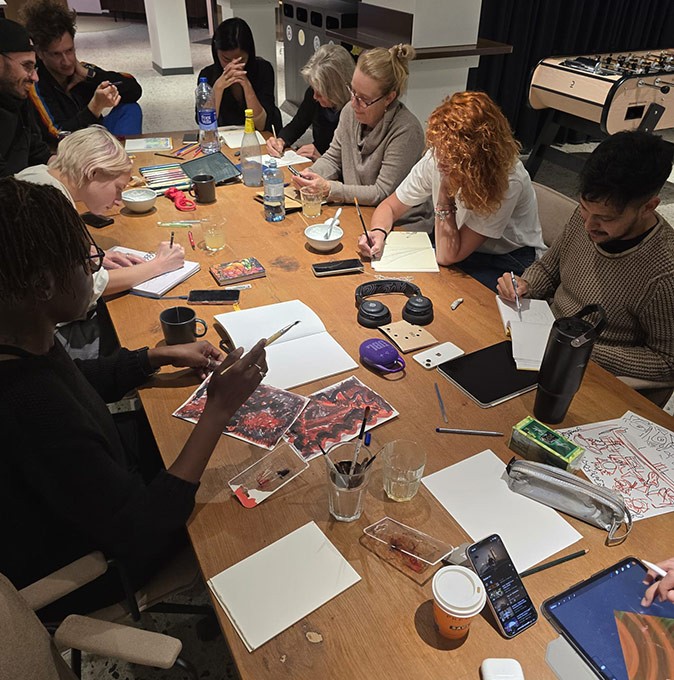 A group of people sitting around a wooden table, working on individual drawings