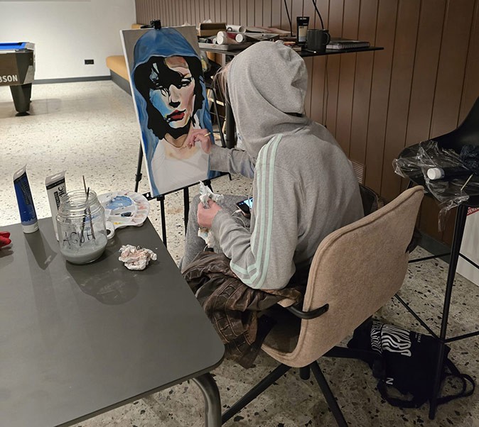 A girl with a gray hoodie sitting on a beige chair, facing a painting of a woman's face she is painting, with painting supplies on a gray table next to her