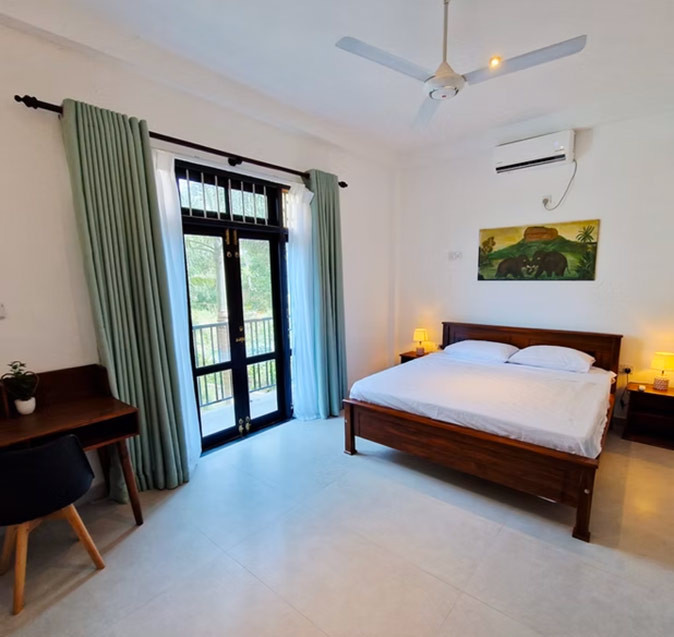 A room at Slow Life Coliving in Sri Lanka A spacious bedroom with a large wooden bed with white sheets, a desk, a window, a painting and air conditioning