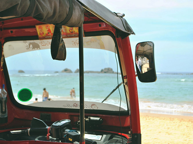 Exploring Sri Lanka while working remotely Part of the interior of a tuktuk, with a sandy beach, a few people and blue sea with some rocks in the background