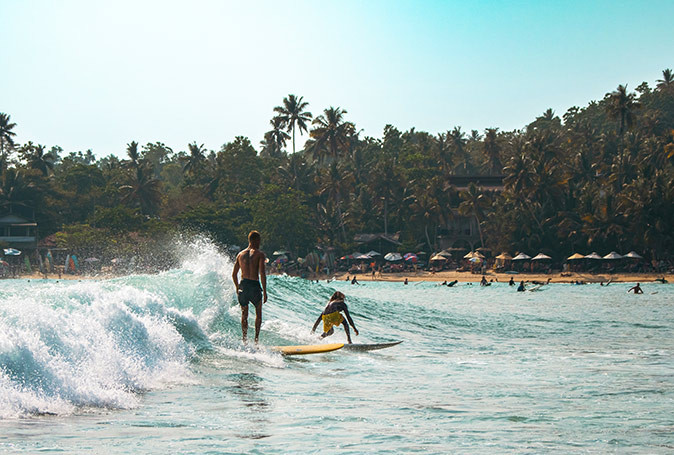 Sri Lanka is a great destination for digital nomads who like to surf 2 surfers riding a wave with a beach with several people, palm trees and umbrellas in the background