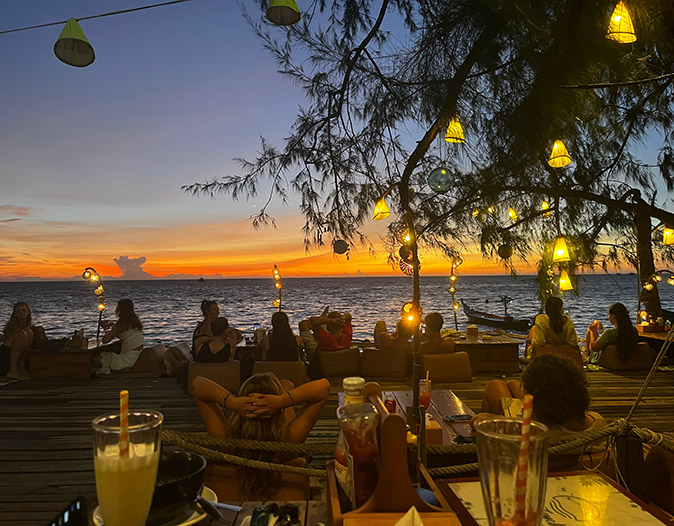 A beach bar showing several people sitting on a wooden deck with the sea and sunset in the background