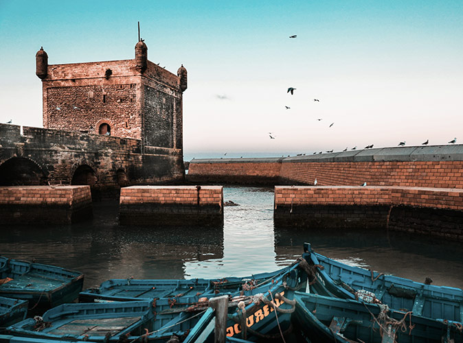 Living in Essaouira, Morocco Part of a historic brown fortification with a bridge and a wall, with pigeons sitting on the wall and flying above it, and several blue wooden boats in the water in front of it