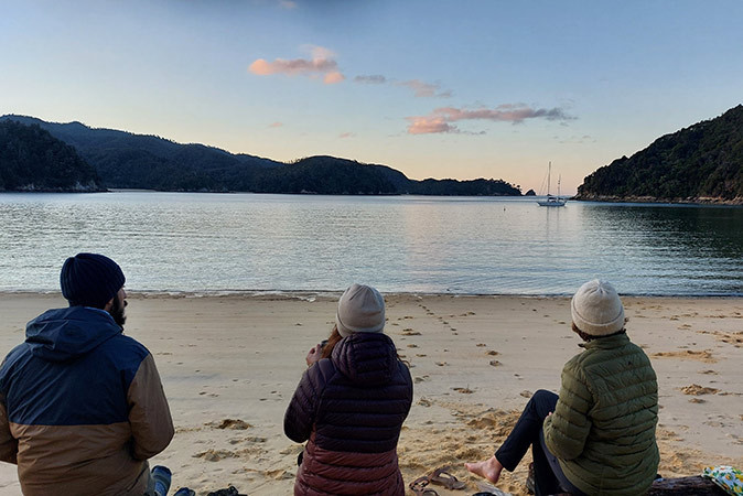 3 people sitting on a sandy beacu looking towards the water with a sailboat and some mountains in the background