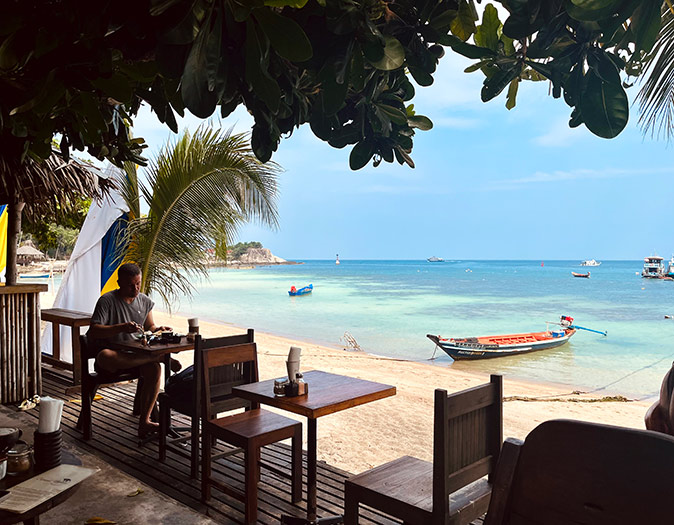 Wooden chairs and tables on a wooden deck with white sand, some colorful boats and crystal clear water in the background