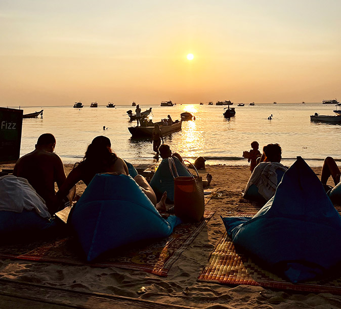 People sitting on bean bags on a sandy beach while the sun sets over a calm sea with several anchored small boats