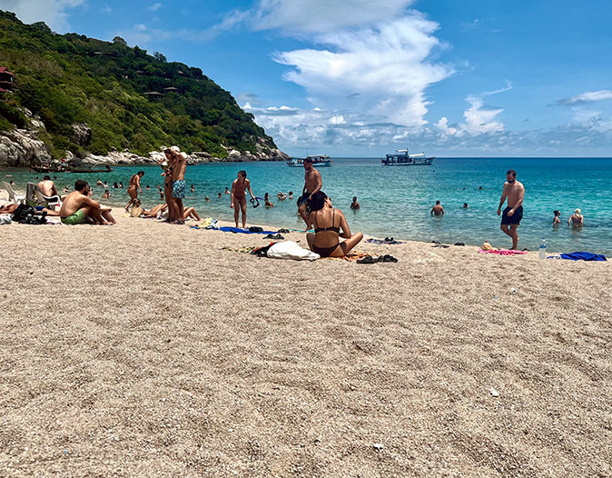 A sandy beach with several people in bathing suits, with a bright blue sea and part of a green hill in the background