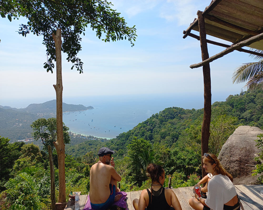 3 people sitting on a wooden platform overlooking a tropical coastline with palmtrees, green hills and bright blue water