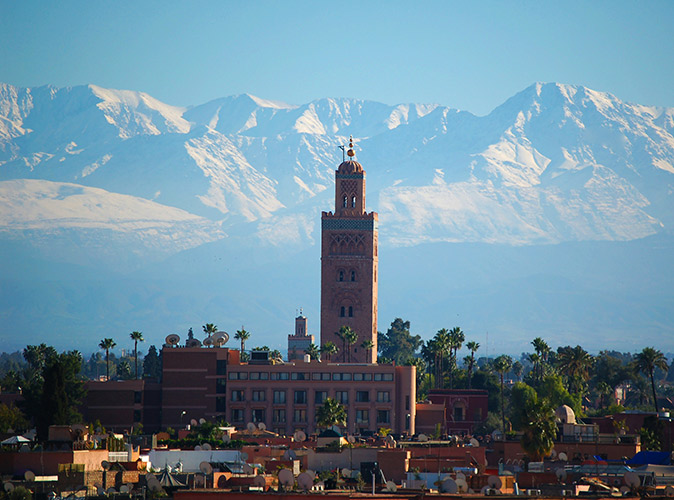 Things to know about living in Morocco Aerial view of Marrakesh showing brown buildings and a large mosque, plus snow-capped mountains in the background
