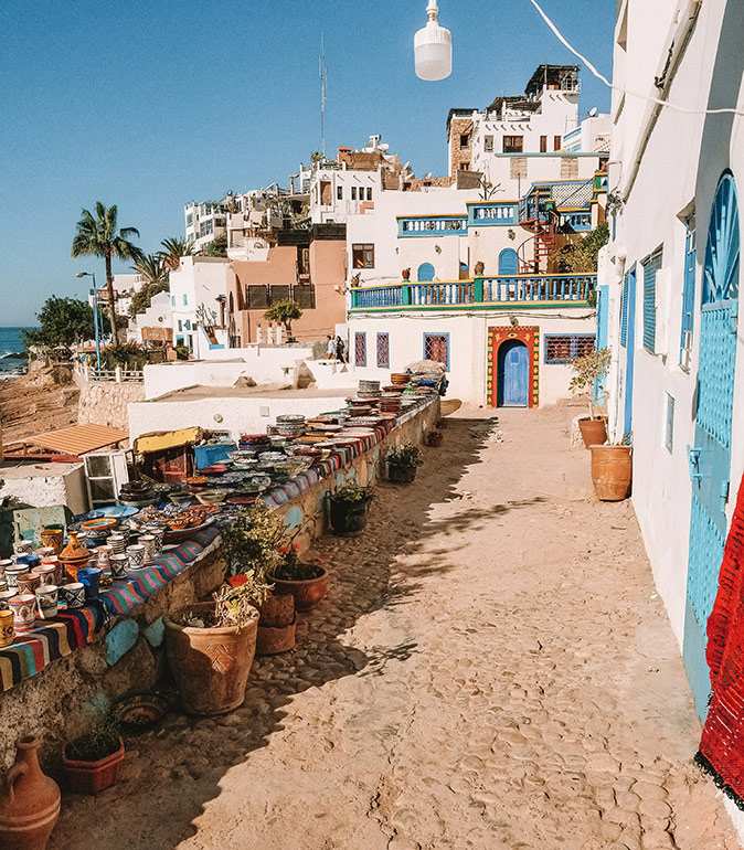 Life in Taghazout, Morocco A typical Moroccan coastal town showing a narrow street with several small items displayed for sale on the left and white and blue buildings on the right and in the background, plus a small part of the beach and ocean