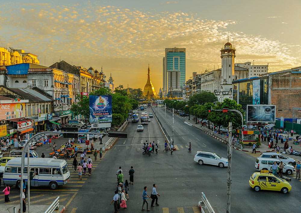 Aerial view of a southeast asian city with a wide road with cars and pedestrians crossing, surrounded by highrise buildings and a gold pagoda showing in the background. Picture taken during susnet
