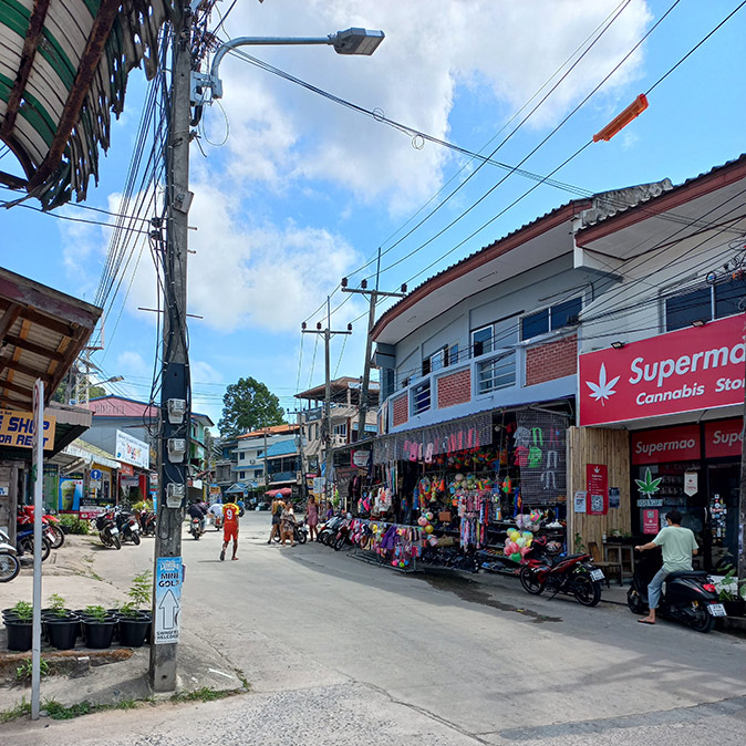 A road with some pedestrians and scooters, lamp posts and 2 story buildings with shops