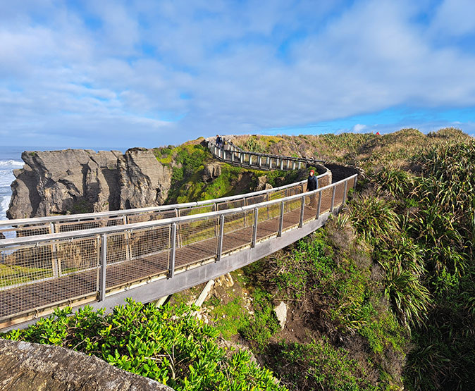 A person walking along a long, metal and wooden bridge, surrounded by greenery and high cliffs