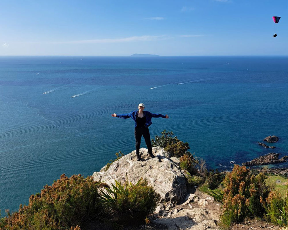 A woman standing on a rock with her arms spread, smiling, with the ocean in the background and a paraglider