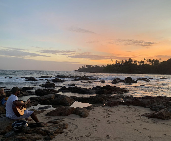 Sri Lanka for digital nomads A sandy beach with several large rocks at sunset showing palm trees in the background and a man with a guitar in the corner