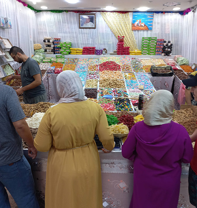 Shopping at a market in Morocco Two women with headscarves facing a market stall with a wide selection of sweets and a man in a tshirt serving them