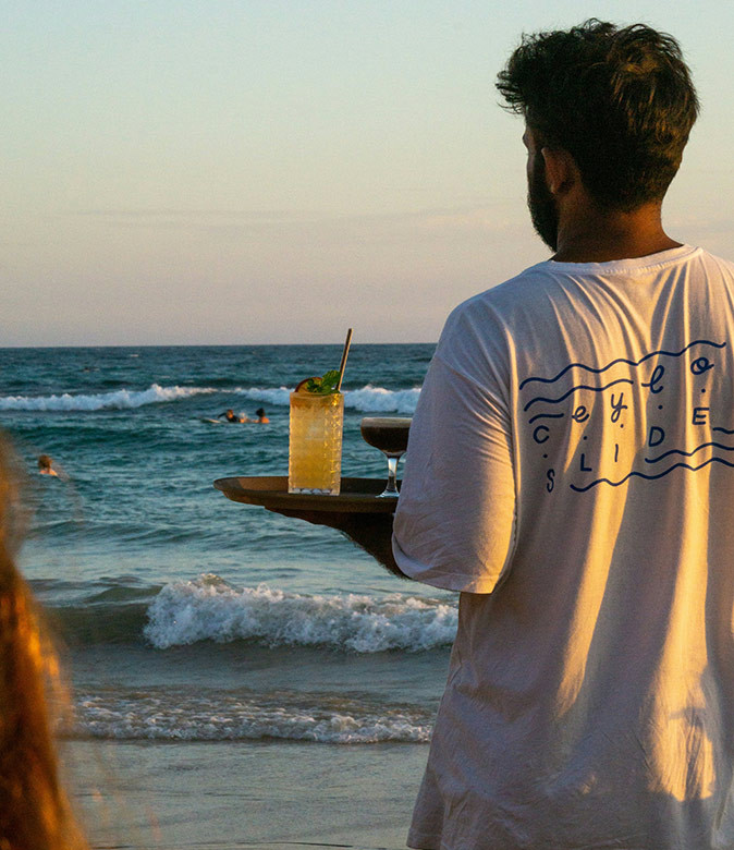 Enjoying after work drinks at Ahangama Beach with digital nomads A man holding a tray with 2 cocktails on a sandy beach with waves in the background