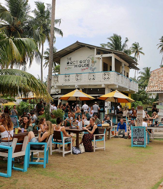 Working remotely from Sri Lanka A white building housing a restaurant with lots of small tables and chairs in front of it filled with people, plus some umbrellas and palm trees