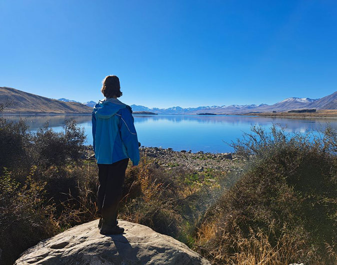 A woman in a blue coat looking out over a large lake surrounde by shrubs
