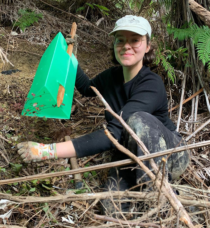 A woman smiling, sitting in a forest surrounded by branches, holding a stick to plant a new tree