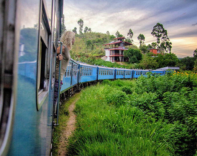 Public transport in Sri Lanka A blue train going through green fields with a blond woman partially hanging out of one of the doors