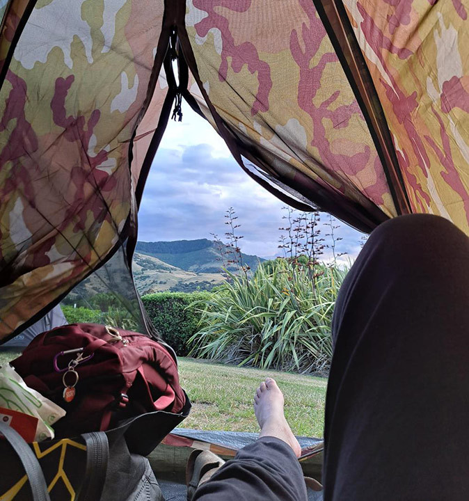 Picture taken from inside a tent showing 2 legs, some camping gear and a view of green hills and a cloudy sky