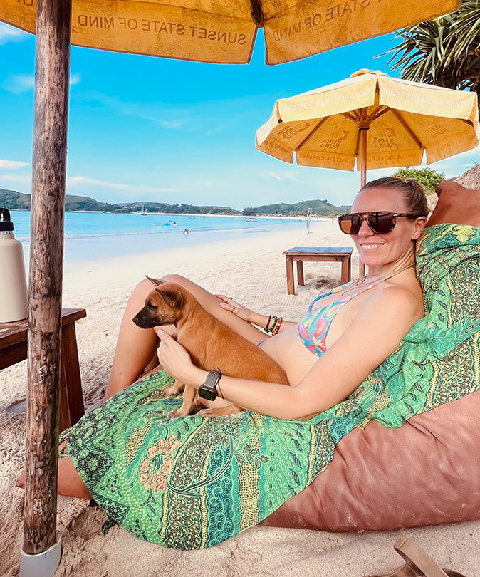 Expat life in Lombok A woman and a dog sitting on a beach under an umbrella with another yellow umbrella in the background plus bright blue water, white sand and some green hills