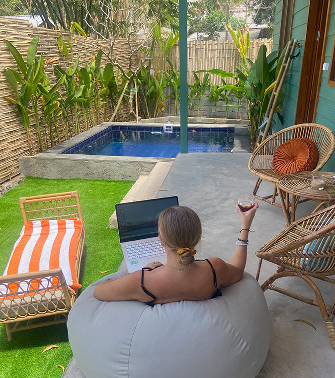 Life in Lombok while working remotely A woman sitting on a bean bag with her laptop and a coffee with a small pool, chairs and some greenery in the background
