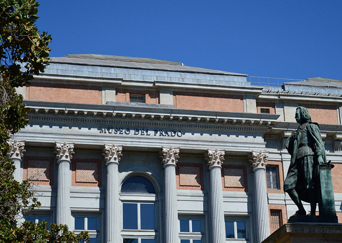 Things to do in Madrid The exterior of the Prado Museum in Madrid, showing a building with gray columns, many windows and a gray and brown exterior, plus a bronze statueof a man in front of it