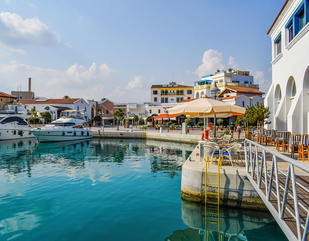 A marina lined with shops and restaurants, some apartment blocks in the background and two luxury yachts in the water