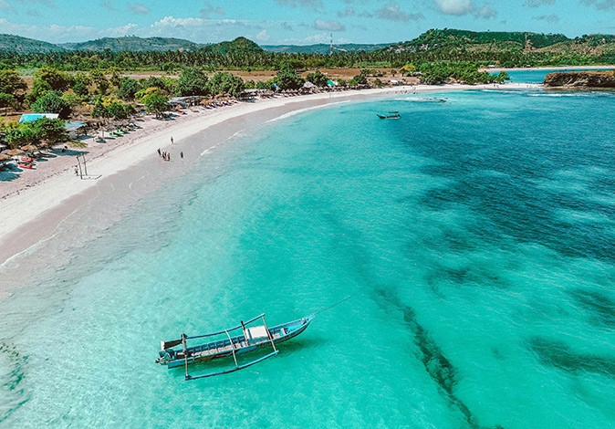 Lombok, Indonesia Aerial view of a tropical island showing greenery, a long sandy beach, bright blue water and a traditional wooden fishing boat
