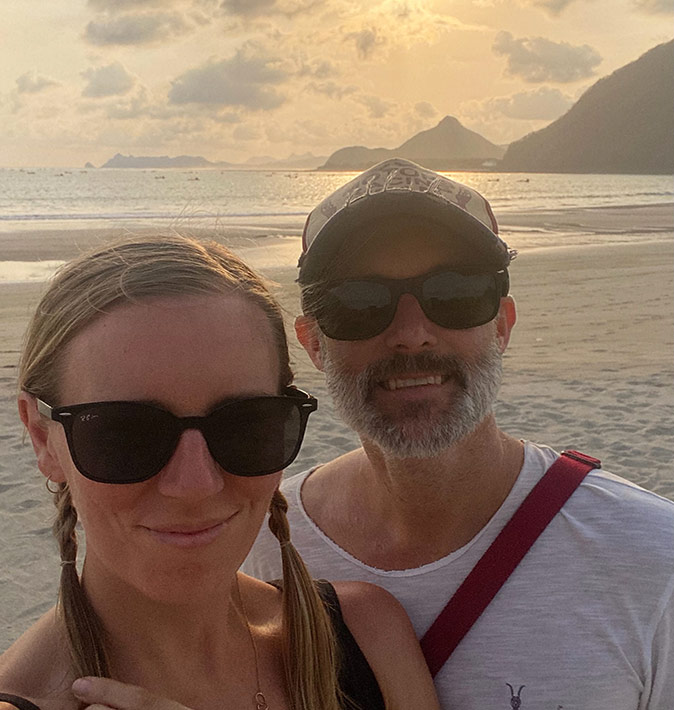 Expats living in Lombok Selfie of a man and a woman with sunglasses, with a sandy beach, sunset and some mountains in the background