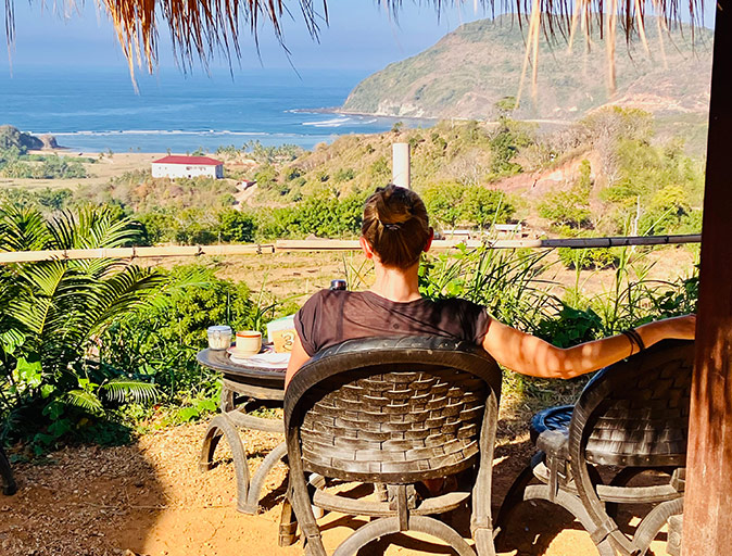 Lombok for expats A woman sitting on a wooden chair looking towards a high anlge view of a coastline with green hills and bright blue water